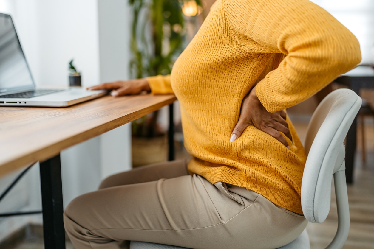 Person holding their lower back in pain while seated at a desk, a possible symptom of a spinal cyst or nerve compression.
